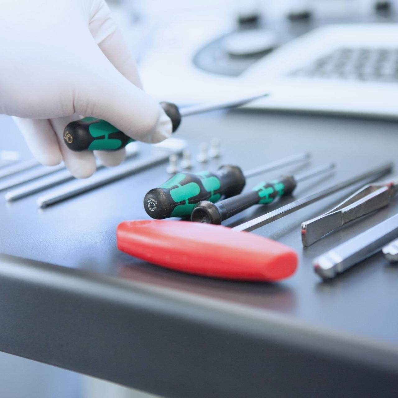 A gloved hand picks up a green-handled screwdriver from a set of precision tools arranged on a black surface. The tools include screwdrivers, tweezers, and other instruments, indicating a repair or maintenance setup.