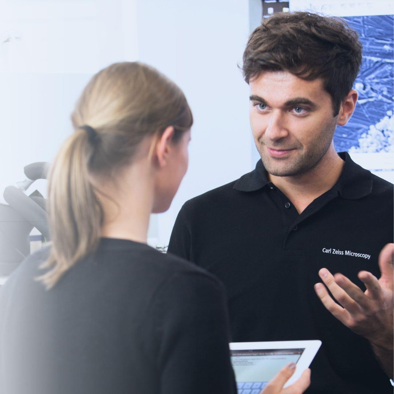A man wearing a Carl Zeiss Microscopy shirt interacts with a woman holding a tablet. The conversation suggests a technical consultation or customer support in a professional setting.