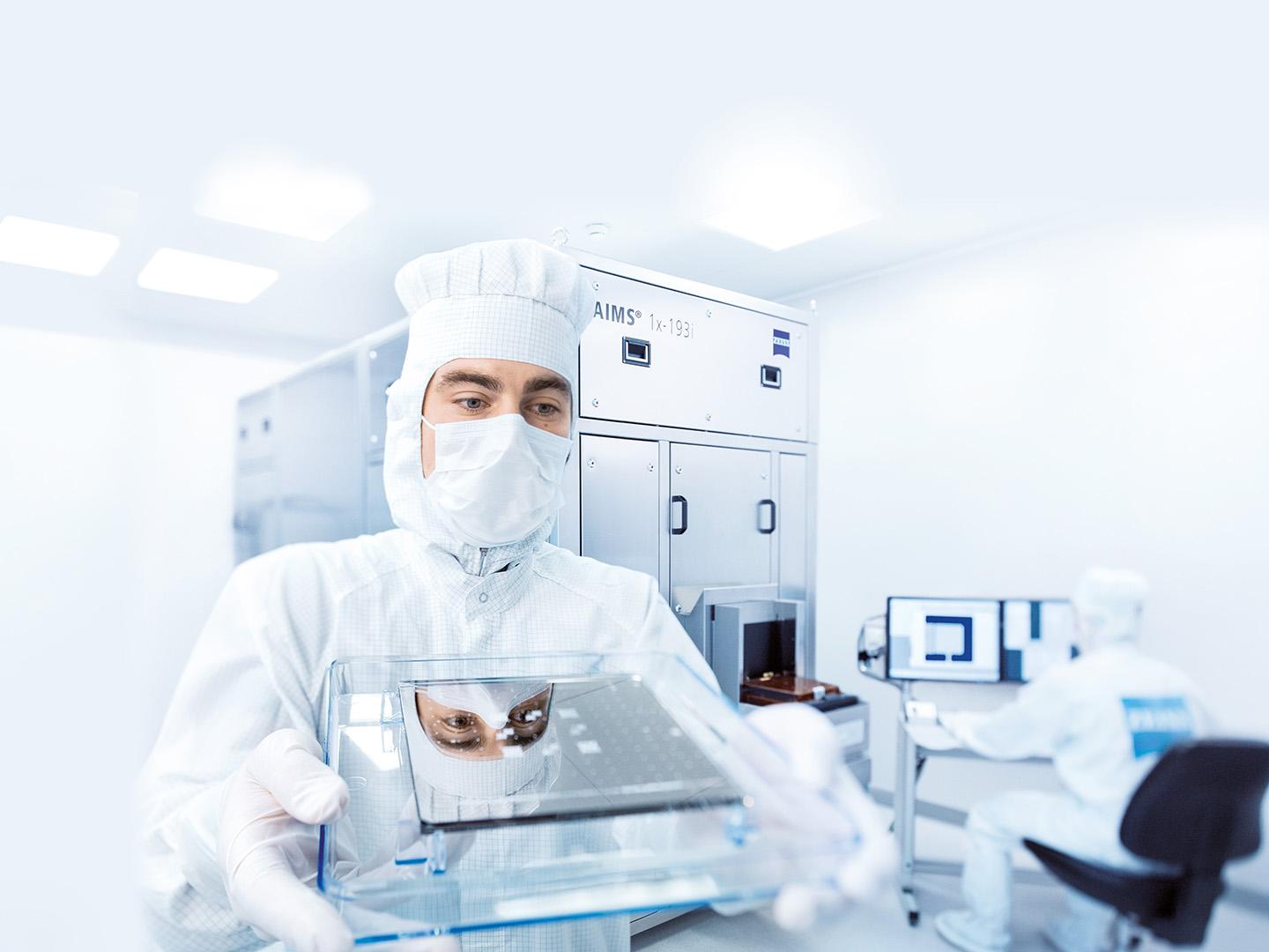 A man in a cleanroom suit holds a photomask in front of him and is reflected in it 