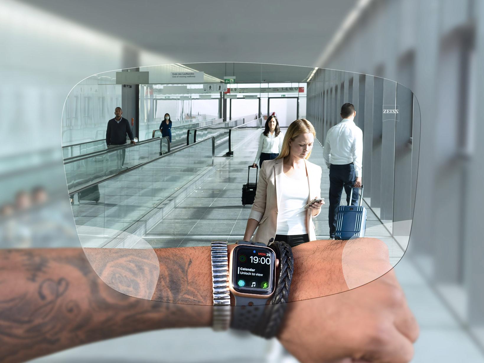 Person looks at his watch through ZEISS SmartLife Digital lenses. In the background you see a typical airport situation with people on escalators pushing their suitcases. 