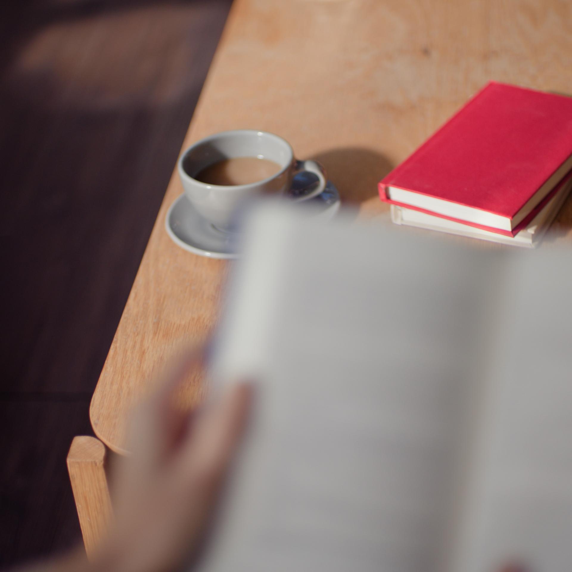 Blurry book held in the hand above a wooden table with a cup of coffee and 2 books on it.