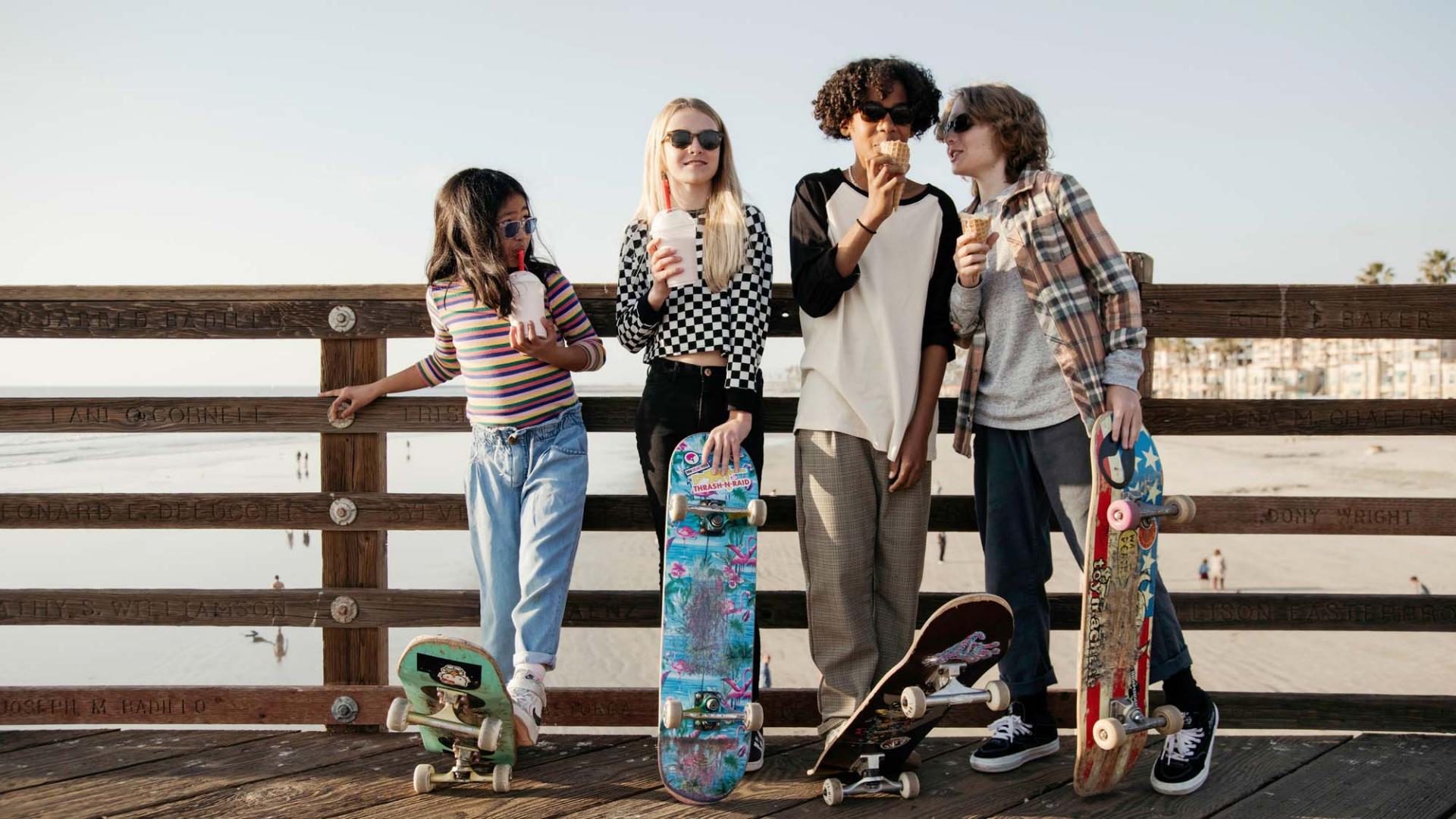 A group of four kids and children standing together. They wear sunglasses drink milkshakes and eat ice cream. All four have skateboards.