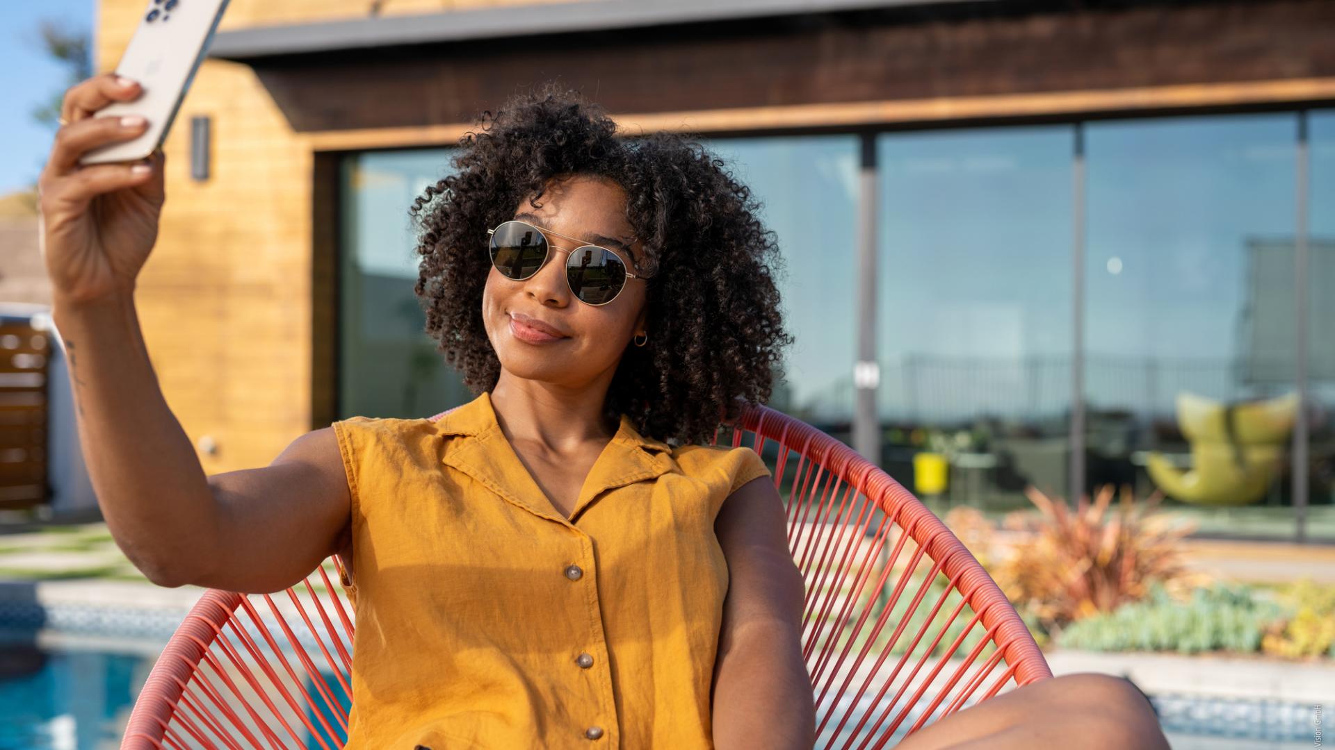 A person with sunglasses sits on a chair in front of a pool and takes a selfie with a smartphone. The colors are bright and cheerful, it is warm.