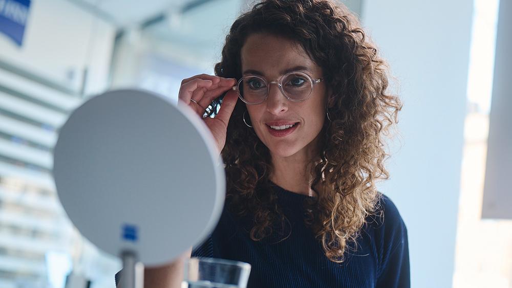 Woman sitting in a ZEISS VISION CENTER in front of a mirror testing new spectacles