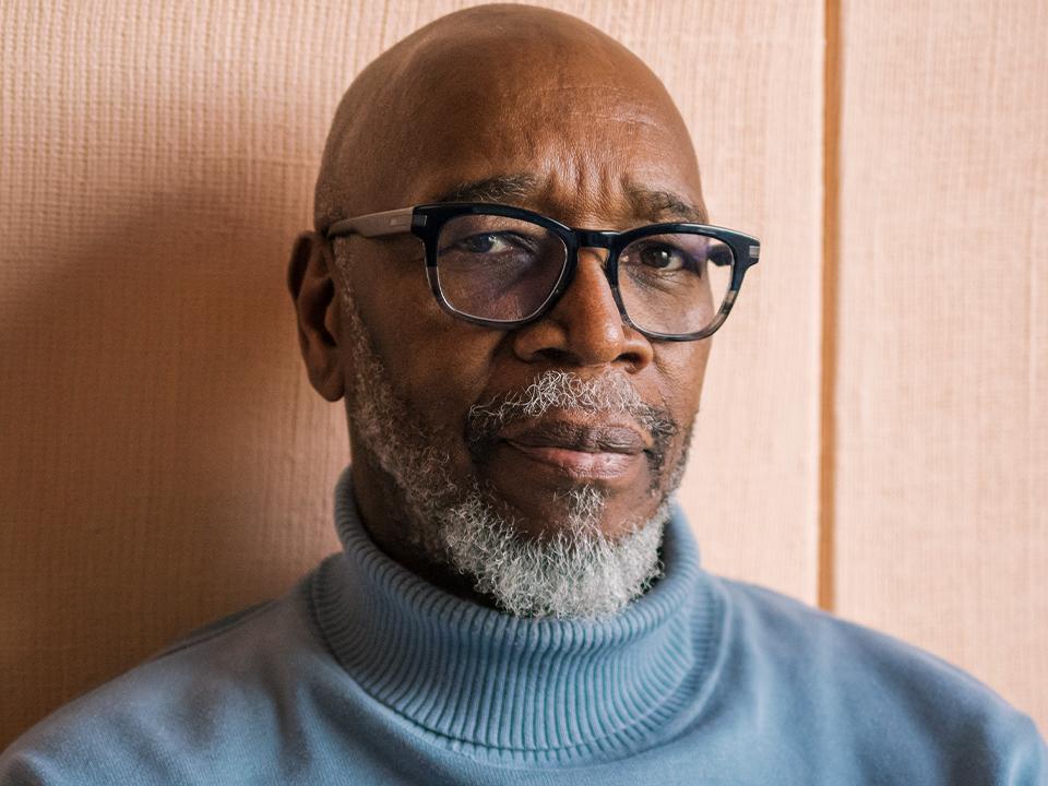 Older man with black glasses and a gray beard looks skeptical. He is wearing a blue turtleneck sweater