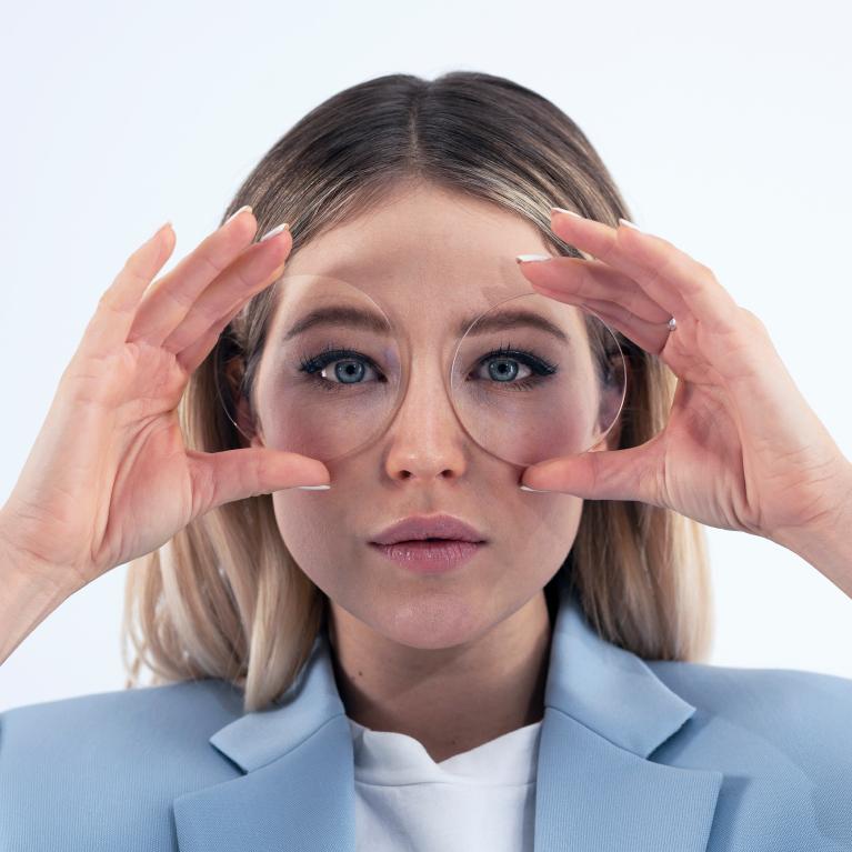 A young blonde woman holds lenses in front of her eyes to show good looks without any funny eye effect thanks to ZEISS Single Vision ClearView lenses.