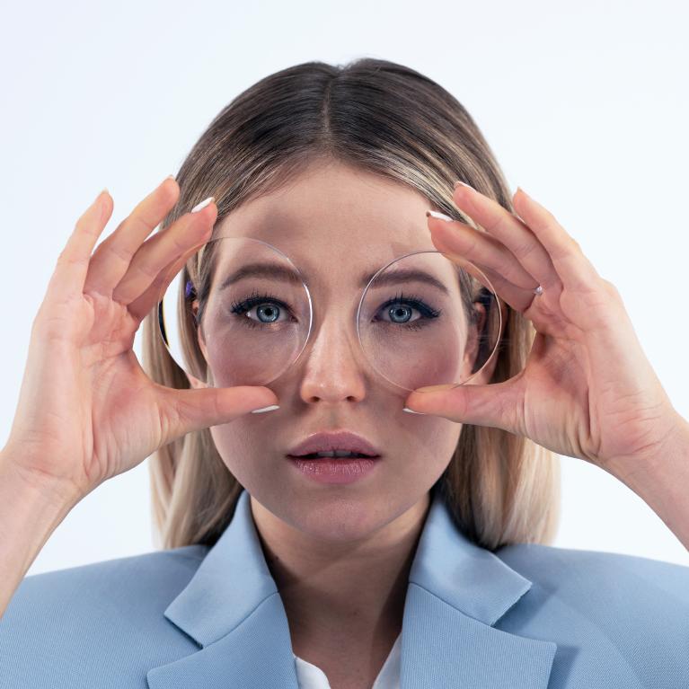 Eine junge blonde Frau hält sich eine Brille vor die Augen und zeigt den von dicken Minusgläsern verursachten Effekt, der die Augen stark verkleinert erscheinen lässt.