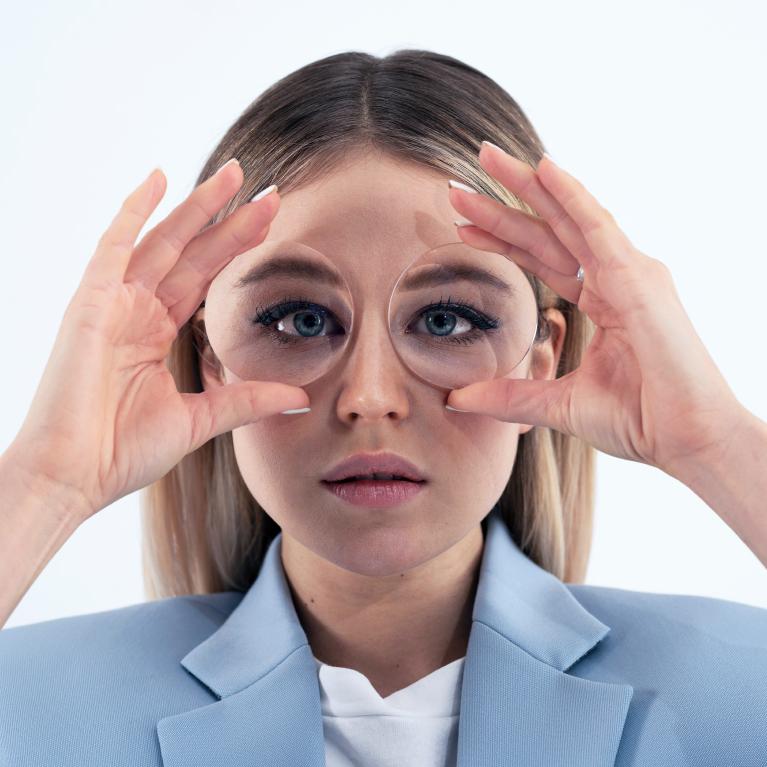 A young blonde woman holds lenses in front of her eyes to show the fish-eye effect caused by thick glasses.