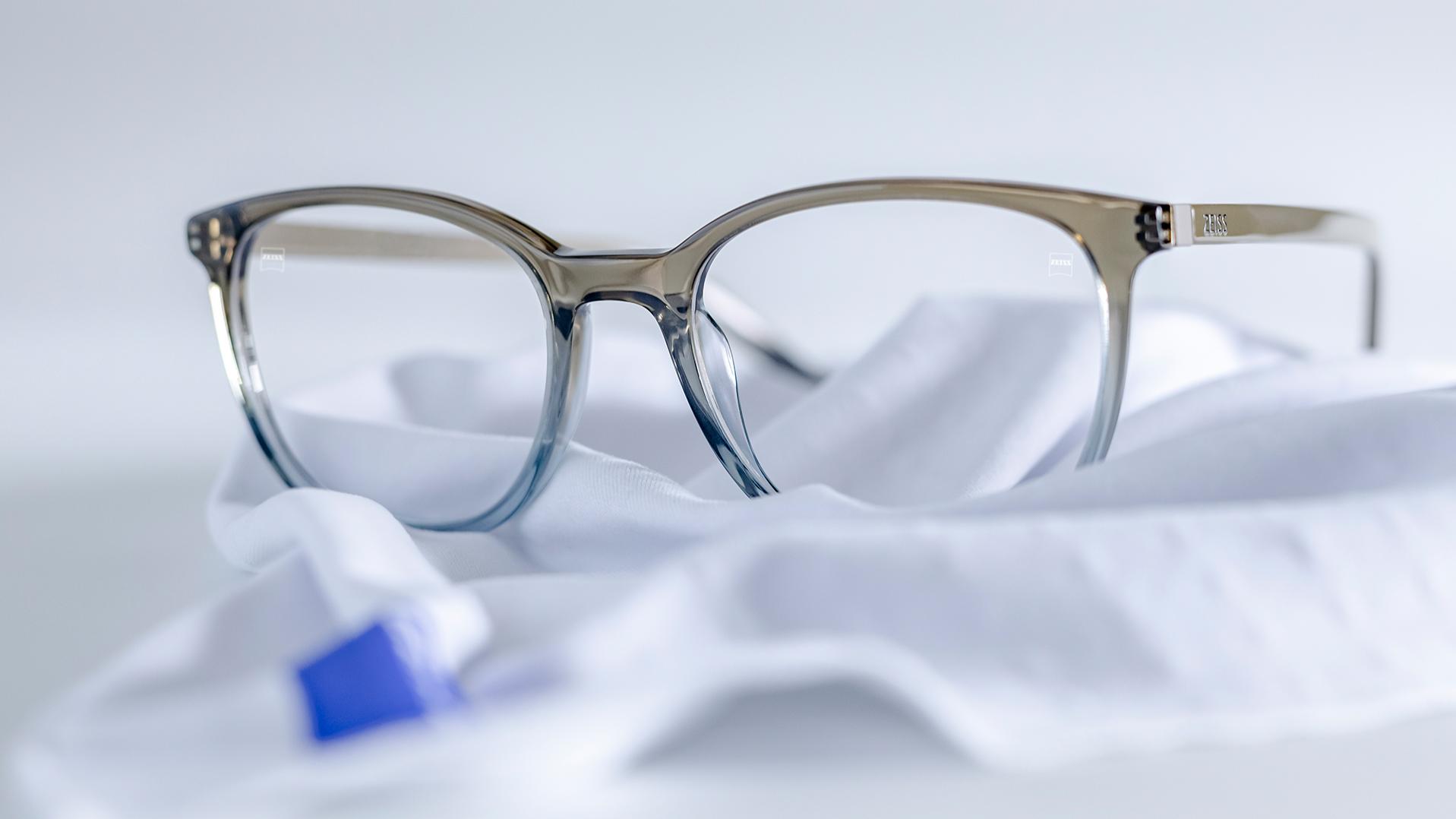 A pair of glasses with gray-blue frames and ZEISS lenses with DuraVision® coating lies on a white microfiber cloth.