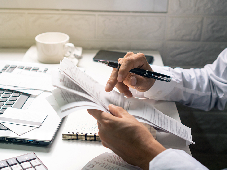 Typical office scene in which a person is busy with reviewing spreadsheets and documents. Only the arms of the person are visible, holding a stack of spreadsheets in his hands. In the background on the desk is a laptop. At the top left of the image is a pop up which shows a male person. The person has gray hair and beard, wears ZEISS Office lenses and has a neutral facial expression.