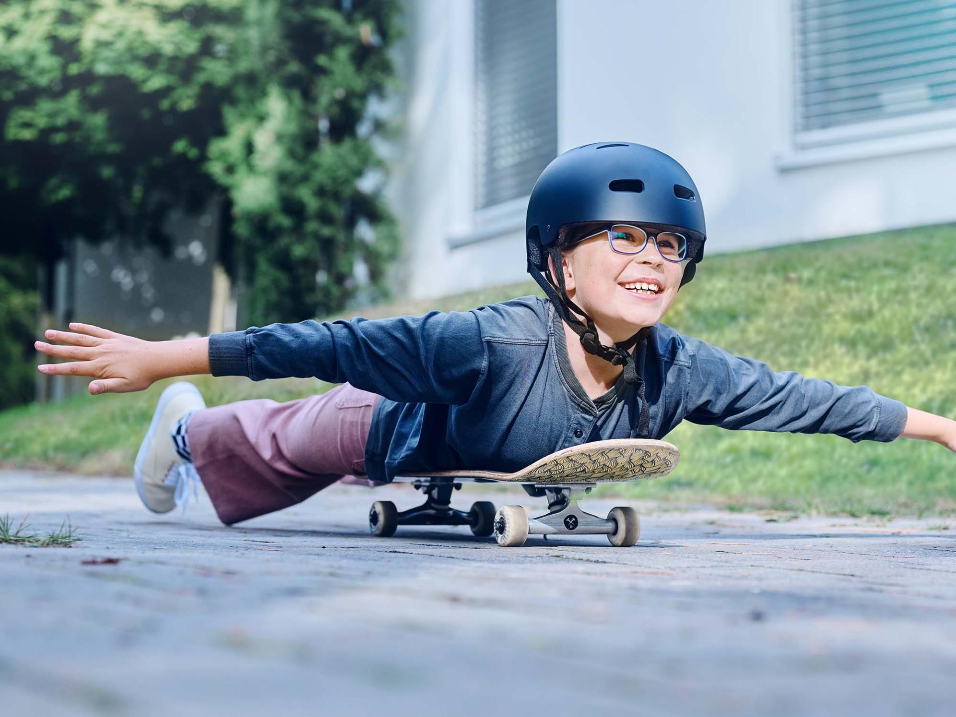 A girl with a helmet and glasses rolls down the road lying on a skateboard with her arms stretched out.