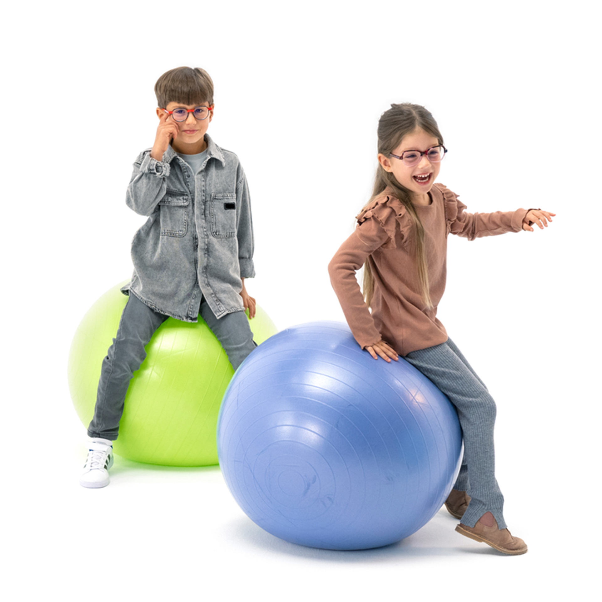A boy and a girl, both wearing glasses, are bouncing playfully on gym balls.