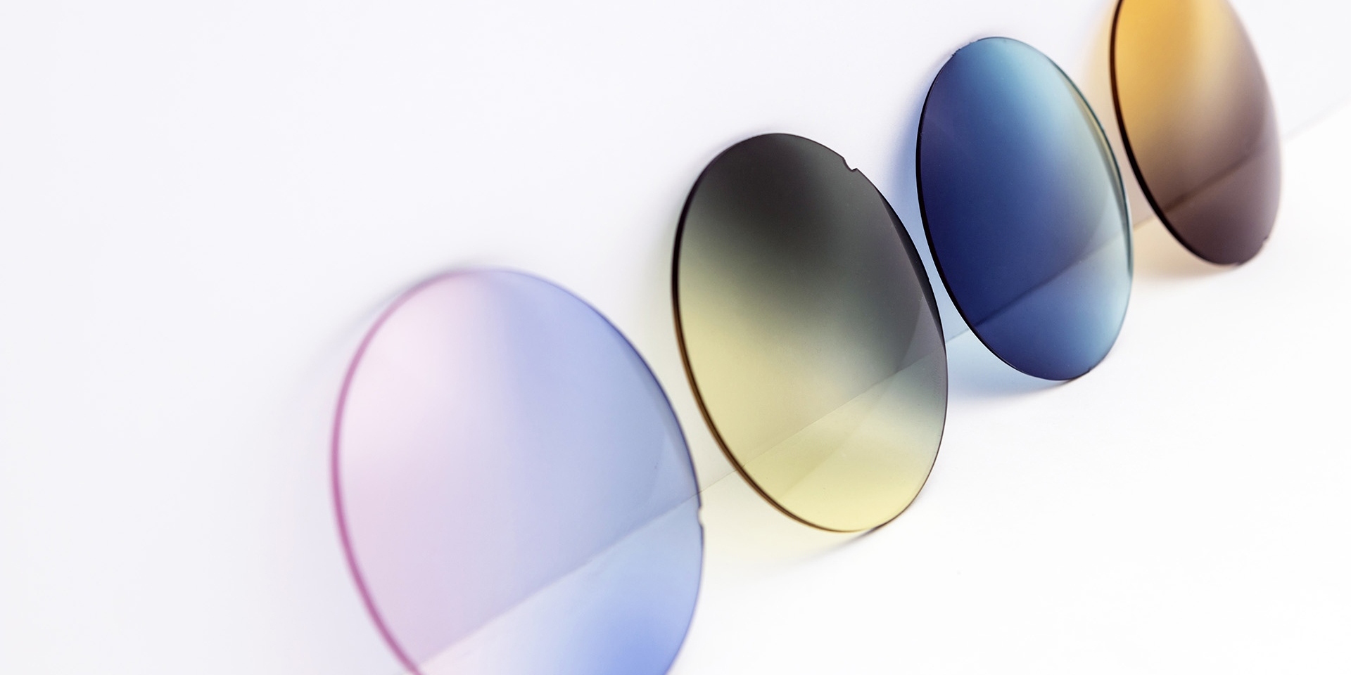 Different colored sunglass lenses leaning against a white surface: pink-purple, yellow-gray, blue and brown gradients.