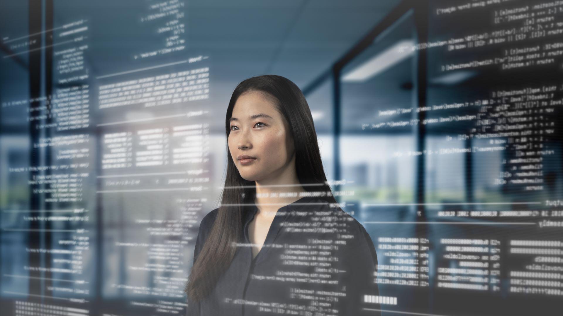A woman stands in a modern office environment with transparent digital code and data projections superimposed in front of her, representing ZEISS technology.