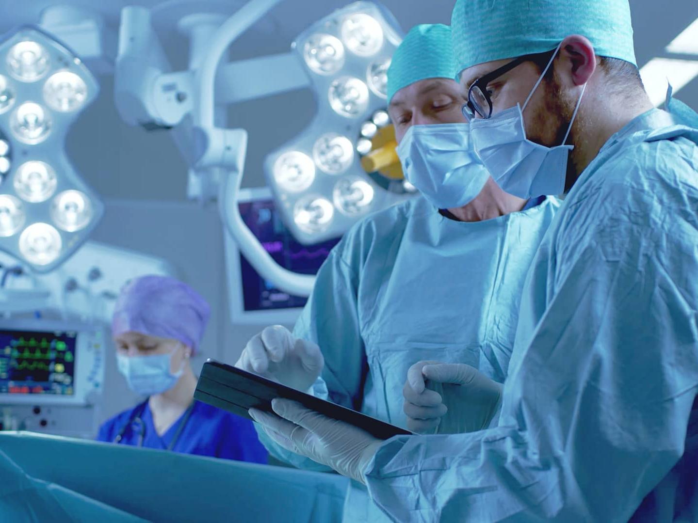 Professional Surgeons and Assistants Talk and Use Digital Tablet Computer while Standing in the Modern Hospital Operating Room.