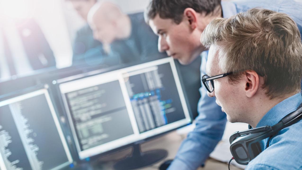 Two software developers are collaborating while analyzing code on multiple computer monitors in a modern office environment.