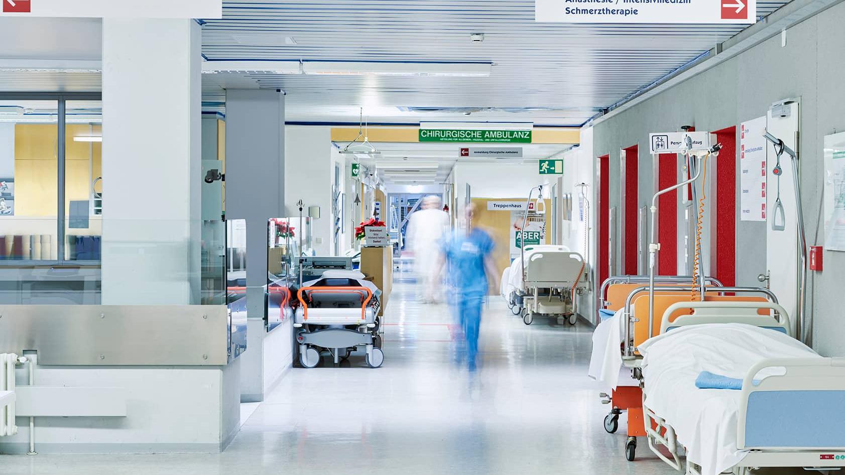 A hospital corridor with medical beds, equipment, and a blurred figure of a healthcare professional walking.